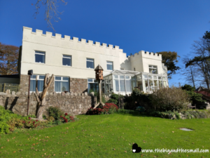 A large white house with a stone base features a castle-like design with battlements. Perfect as a hotel and commercial building, it offers significant venue and development potential. The front garden is green and well-maintained, with shrubs, flowers, and a birdhouse. A tall tree front left and blue sky complete the scene. Website text is in the bottom right corner.