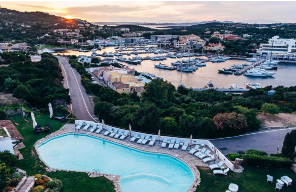 An aerial view of a coastal resort at sunset, featuring a large, crystal-clear swimming pool surrounded by lounge chairs and umbrellas. The pool area overlooks a marina filled with boats, lush greenery, and a winding road. In the background, hills and buildings are illuminated by the setting sun.