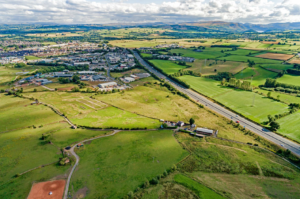 Aerial view of a verdant countryside with a mix of green fields, patches of forests, and scattered buildings. A highway runs diagonally through the landscape. Rolling hills and distant mountains are visible under a partly cloudy sky. This picturesque land is for sale with Palace Auctions.