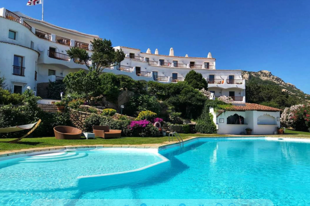 A beautiful pool area at a luxury resort, surrounded by lush greenery and colorful flowers. A multi-story white building with balconies overlooks the pool. There are lounge chairs, a hammock, and a bright blue sky above. Mountains are visible in the background.