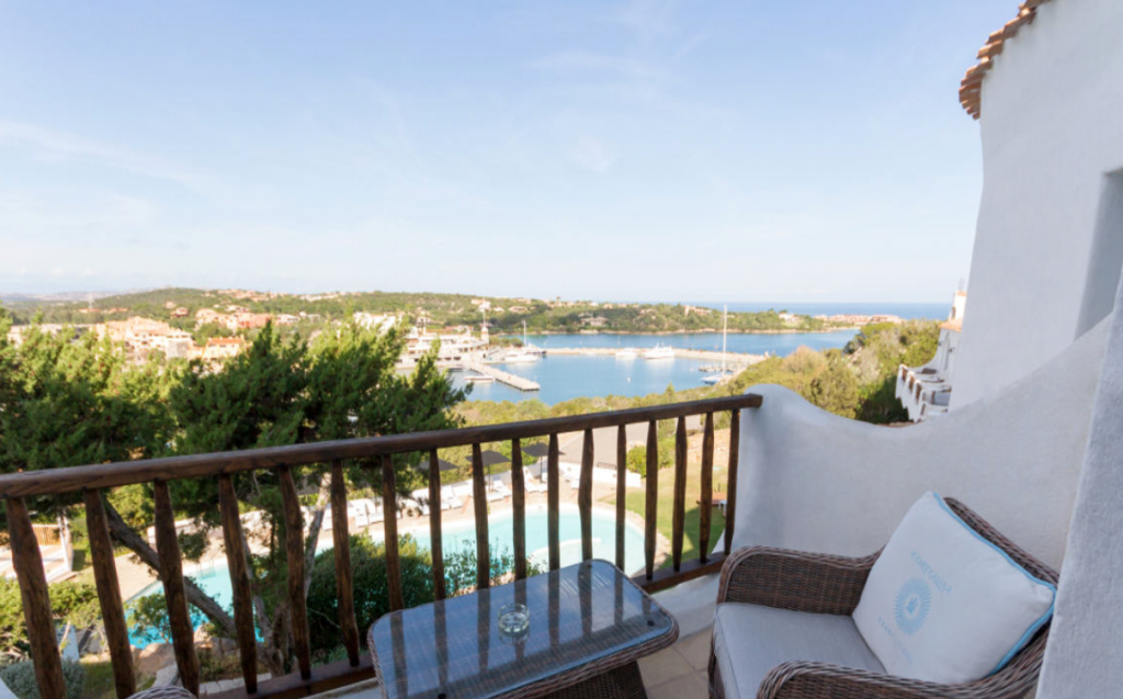 A scenic view of a coastal area from a balcony with a wooden railing. The balcony has a couple of wicker chairs with cushions and a small glass table. Below is a round swimming pool surrounded by greenery. Beyond is a marina with boats and a hilly landscape.