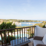 A scenic view of a coastal area from a balcony with a wooden railing. The balcony has a couple of wicker chairs with cushions and a small glass table. Below is a round swimming pool surrounded by greenery. Beyond is a marina with boats and a hilly landscape.