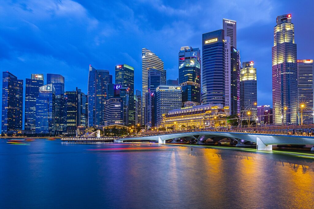 A nighttime cityscape of Singapore's Marina Bay, featuring numerous illuminated skyscrapers reflecting on the water. A bridge spans the bay on the right, with blurred lights of passing boats creating colorful trails across the calm blue waters—a picturesque setting for an Office Investment Business for Sale in Singapore.
