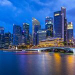 A nighttime cityscape of Singapore's Marina Bay, featuring numerous illuminated skyscrapers reflecting on the water. A bridge spans the bay on the right, with blurred lights of passing boats creating colorful trails across the calm blue waters—a picturesque setting for an Office Investment Business for Sale in Singapore.