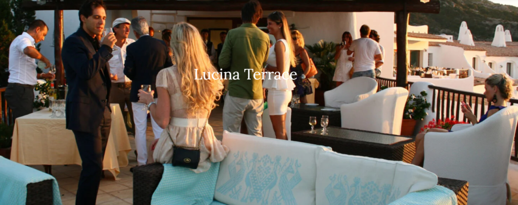 People are socializing on the Lucina Terrace. The outdoor space features modern wicker furniture with light cushions, and tables are set with white tablecloths. The background includes a scenic hillside and a glimpse of white buildings.