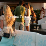 People are socializing on the Lucina Terrace. The outdoor space features modern wicker furniture with light cushions, and tables are set with white tablecloths. The background includes a scenic hillside and a glimpse of white buildings.