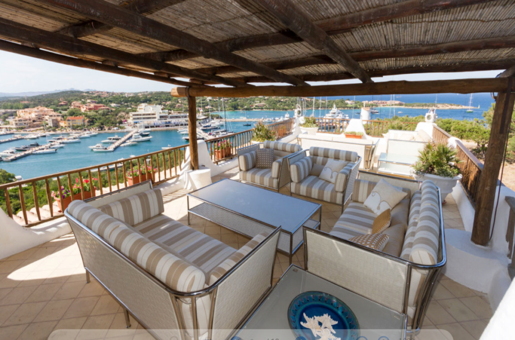 An outdoor patio area with striped cushioned seating, glass-topped coffee tables, and a pergola overhead. The view overlooks a marina with numerous boats, calm blue waters, and a coastal town in the background under a clear sky.