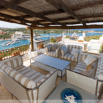 An outdoor patio area with striped cushioned seating, glass-topped coffee tables, and a pergola overhead. The view overlooks a marina with numerous boats, calm blue waters, and a coastal town in the background under a clear sky.