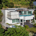 A modern, multi-level white house featuring large windows and a swimming pool on the second floor. The home is surrounded by lush greenery and trees. The design incorporates sleek lines and balconies, set amidst a tropical landscape.