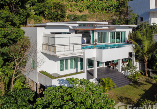 A modern, multi-level white house featuring large windows and a swimming pool on the second floor. The home is surrounded by lush greenery and trees. The design incorporates sleek lines and balconies, set amidst a tropical landscape.