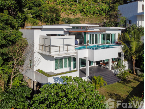 A modern, multi-level white house featuring large windows and a swimming pool on the second floor. The home is surrounded by lush greenery and trees. The design incorporates sleek lines and balconies, set amidst a tropical landscape.
