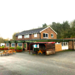 Property for sale by Auction A brick building features signs for "The Barn House" pub, also known as the Three Lions Public House. It has a pitched roof and a single-story extension. In front, picnic tables and wooden benches are arranged on a paved area. Trees and a cloudy sky are in the background. Presented by Palace Auctions