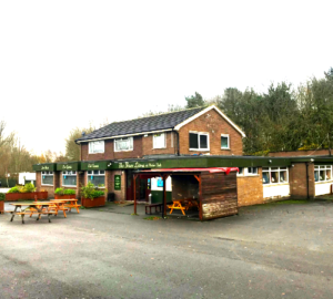 Property for sale by Auction A brick building features signs for "The Barn House" pub, also known as the Three Lions Public House. It has a pitched roof and a single-story extension. In front, picnic tables and wooden benches are arranged on a paved area. Trees and a cloudy sky are in the background. Presented by Palace Auctions