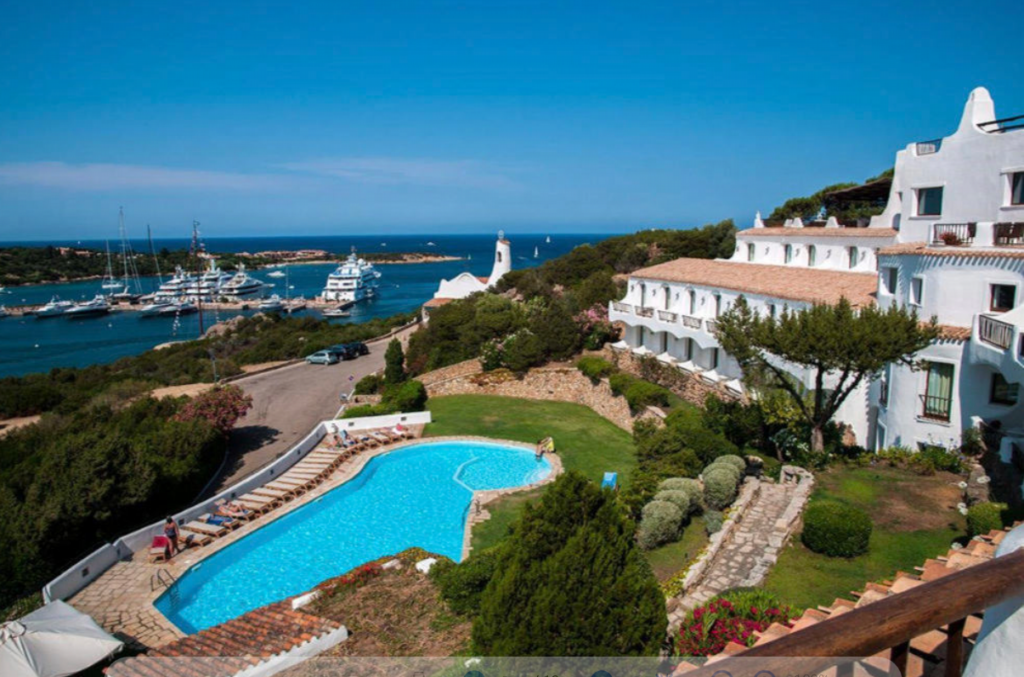 A scenic view of a coastal resort shows a large swimming pool surrounded by lounge chairs and greenery in the foreground. Mediterranean-style white buildings are on the right, and several yachts are docked in a clear blue marina on the left, against a backdrop of the sea.