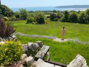 A person in a colorful dress stands on a wooden pathway, facing a lush green field with wildflowers. Beyond the field lies the scenic view of Nicolaston House Gardens, featuring an ocean coastline and blue sky, surrounded by greenery, rocks, and flowers in the foreground.