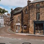 A picturesque street corner in a small town features historic stone buildings including St. James's Church Centre. The scene shows a winding road flanked by quaint shops and houses with rooftops, under a slightly cloudy sky. A tree branch is partially visible.