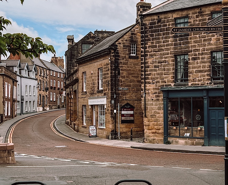 A picturesque street corner in a small town features historic stone buildings including St. James's Church Centre. The scene shows a winding road flanked by quaint shops and houses with rooftops, under a slightly cloudy sky. A tree branch is partially visible.