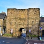 A historic stone gate with a large central archway stands at the entrance of a small town. The gate is flanked by old buildings. Traffic signs and flower beds with bright blooms decorate the area. A road runs through the archway under a clear sky.