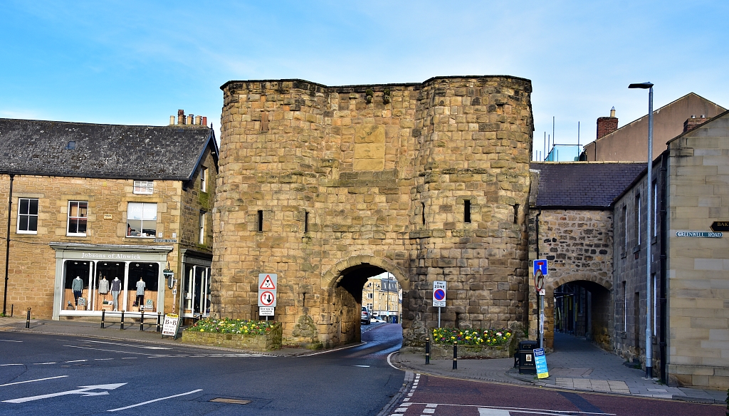 A historic stone gate with a large central archway stands at the entrance of a small town. The gate is flanked by old buildings. Traffic signs and flower beds with bright blooms decorate the area. A road runs through the archway under a clear sky.