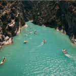 A narrow, turquoise river flows between steep, rocky cliffs covered in greenery. Several kayaks and small boats with people are scattered along the serene waterway. The sunny, picturesque scene, reminiscent of Remi's investment opportunities in Phuket, features a clear blue sky above.