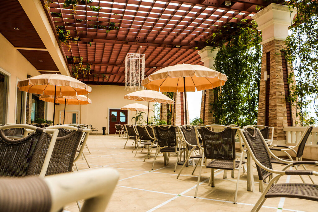 A spacious outdoor dining area with several tables and chairs under large beige umbrellas. A pergola with greenery and hanging decorations provides partial shade. The serene setting is ideal for relaxed dining at the Grand Pacific Sovereign Resort & Spa, an off-market hotel investment opportunity.
