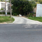 A small side street branches off from a main road, curving to the left. The side street is surrounded by greenery, including trees and bushes. A utility pole and building with a gray wall are on the left, and a sheet metal fence is on the right.