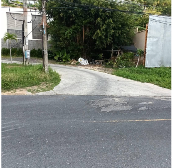 A small side street branches off from a main road, curving to the left. The side street is surrounded by greenery, including trees and bushes. A utility pole and building with a gray wall are on the left, and a sheet metal fence is on the right.