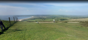 A scenic view of a grassy hilltop overlooking a coastal landscape. Near the center, a person stands looking towards the ocean with a dog nearby. In the distance, Nicholaston House in Swansea adds charm to the sandy beach curving along the coastline, bounded by lush green fields under a clear blue sky.