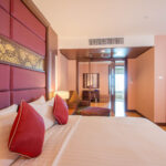 A spacious hotel room at the Grand Pacific Sovereign Resort & Spa features a large bed adorned with red pillows and a gold-patterned headboard. To the left, there's a seating area with patterned cushions. The room leads to an adjoining space with another bed in the background. The lighting is warm and inviting.