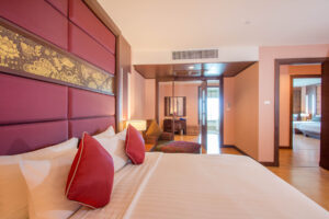 A spacious hotel room at the Grand Pacific Sovereign Resort & Spa features a large bed adorned with red pillows and a gold-patterned headboard. To the left, there's a seating area with patterned cushions. The room leads to an adjoining space with another bed in the background. The lighting is warm and inviting.
