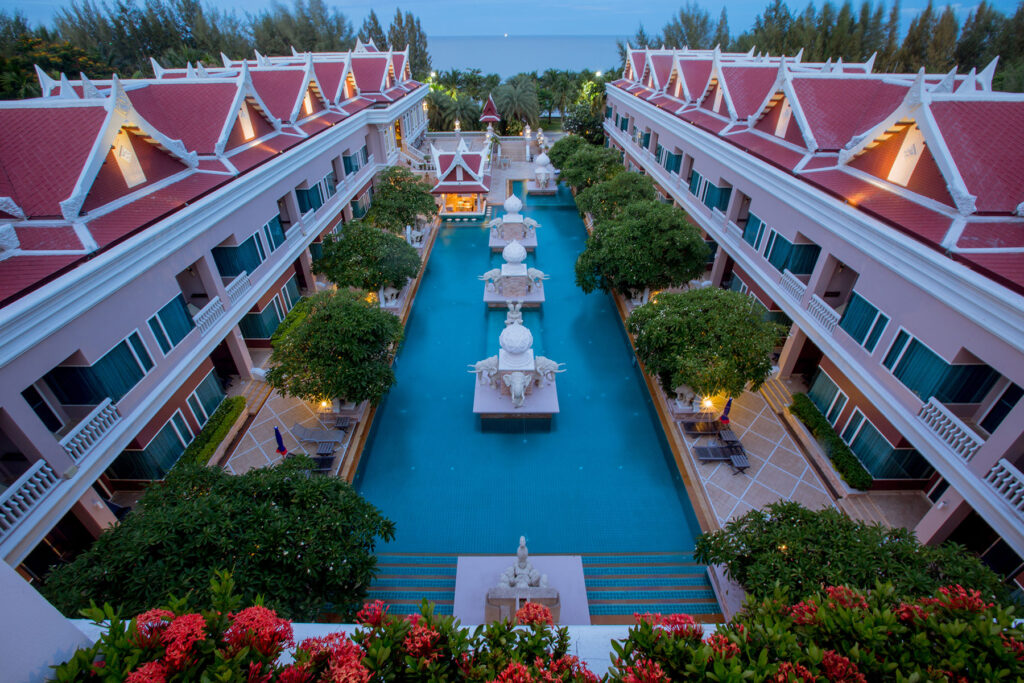 Aerial view of the Grand Pacific Sovereign Resort & Spa featuring a long central swimming pool flanked by two symmetrical, ornate buildings with red roofs. The pool is adorned with decorative statues and surrounded by lush greenery, with the open sea visible in the background.