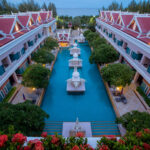 Aerial view of the Grand Pacific Sovereign Resort & Spa featuring a long central swimming pool flanked by two symmetrical, ornate buildings with red roofs. The pool is adorned with decorative statues and surrounded by lush greenery, with the open sea visible in the background.