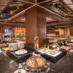 A lavish buffet setup at the Grand Pacific Sovereign Resort & Spa features a variety of dishes on display. Neatly arranged plates, bowls, trays of fruits, eggs, sauces, and a selection of hot and cold foods complement the room's modern decor with wood and glass elements and ample lighting.