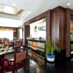 A modern restaurant interior at Grand Pacific Sovereign Resort & Spa features dark wooden decor, tables with cushioned chairs, and a buffet section displaying various foods. Potted plants add greenery, while large windows allow natural light to illuminate this exquisite space.