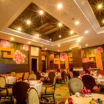 A warmly lit, elegant restaurant interior at the Grand Pacific Sovereign Resort & Spa features floral wallpaper, round tables covered with white cloths, and cushioned chairs. Each table is set with red napkins and dinnerware. Recessed and pendant lighting illuminate the room, creating a cozy ambiance.