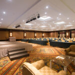 A spacious, well-lit conference room at the Grand Pacific Sovereign Resort & Spa features a carpeted floor, rows of tables with chairs facing a stage, and comfortable armchairs around glass tables at the front. The stage is equipped with a podium and decorated with a floral arrangement.