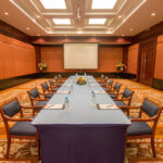 A conference room at the Grand Pacific Sovereign Resort & Spa is set up with a U-shaped table arrangement covered in a blue tablecloth. Chairs with blue seats surround the table and notebooks and water bottles are placed at each seat. At the front is a large projector screen, and floral arrangements are in the corners.