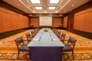A conference room at the Grand Pacific Sovereign Resort & Spa is set up with a U-shaped table arrangement covered in a blue tablecloth. Chairs with blue seats surround the table and notebooks and water bottles are placed at each seat. At the front is a large projector screen, and floral arrangements are in the corners.