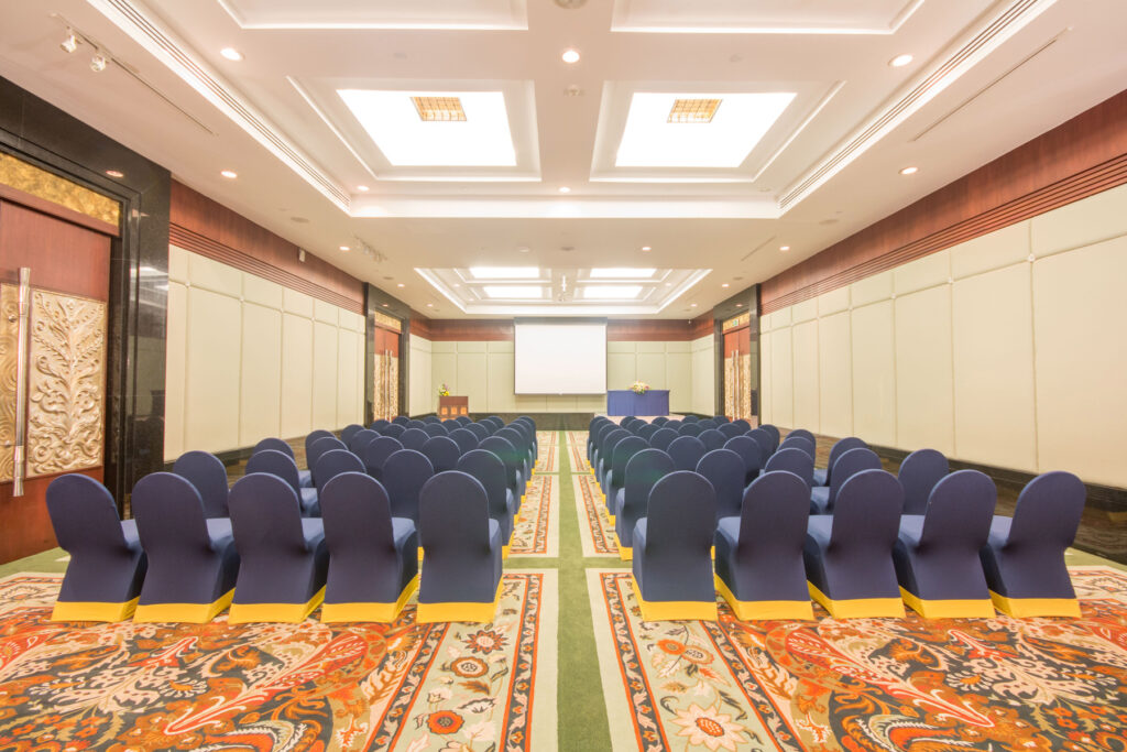 The Grand Pacific Sovereign Resort & Spa offers an off-market hotel investment opportunity. An empty conference room, set up with rows of blue-covered chairs facing a large white screen, is well-lit with ceiling lights and features ornate decorations on the walls and floor, including intricate patterns and a colorful carpet.