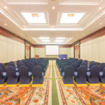 The Grand Pacific Sovereign Resort & Spa offers an off-market hotel investment opportunity. An empty conference room, set up with rows of blue-covered chairs facing a large white screen, is well-lit with ceiling lights and features ornate decorations on the walls and floor, including intricate patterns and a colorful carpet.