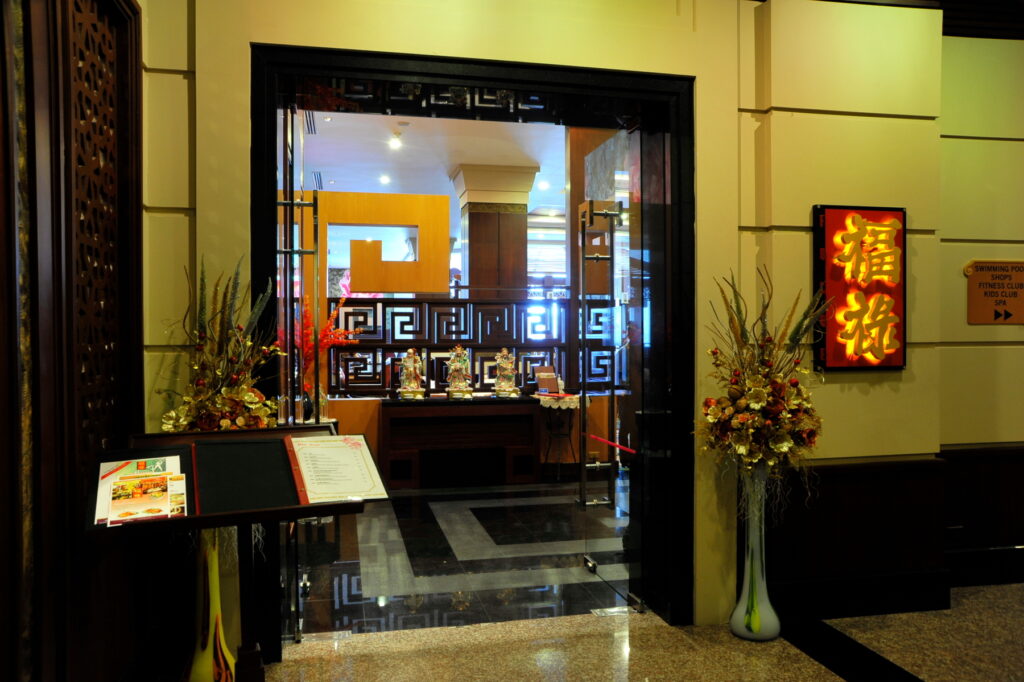 A warmly lit restaurant entrance at the Grand Pacific Sovereign Resort & Spa features a pair of tall decorative vases with flowers on either side of the doorway. Inside, intricate wooden partitions, a buffet display, and a golden wall decoration with red Chinese characters create an inviting atmosphere.