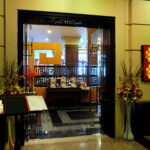 A warmly lit restaurant entrance at the Grand Pacific Sovereign Resort & Spa features a pair of tall decorative vases with flowers on either side of the doorway. Inside, intricate wooden partitions, a buffet display, and a golden wall decoration with red Chinese characters create an inviting atmosphere.