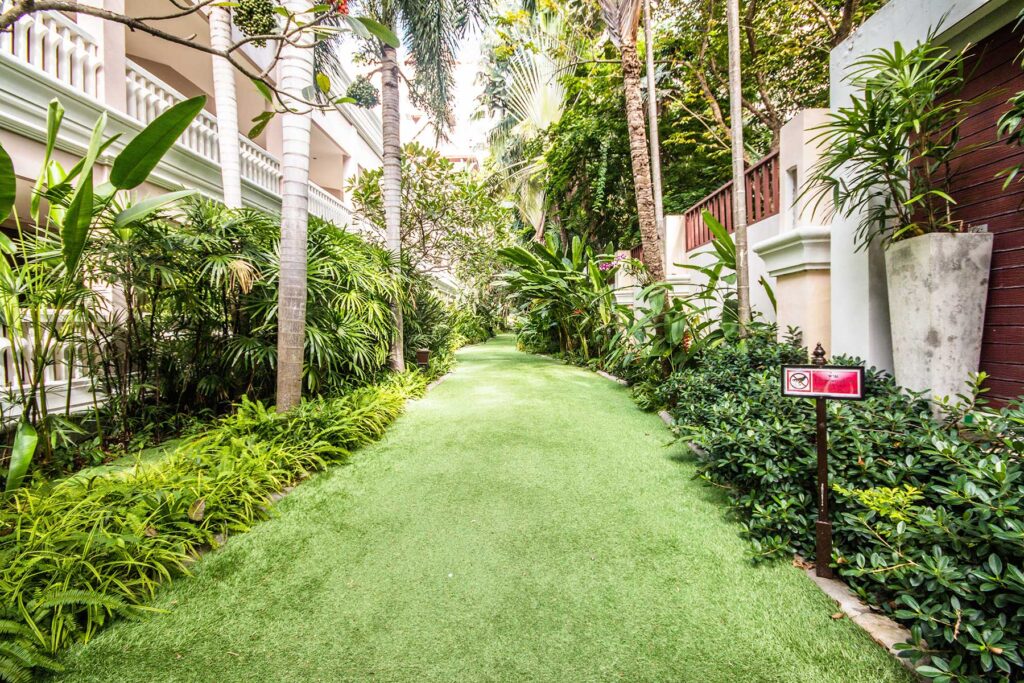 A serene pathway with lush green artificial grass flanked by various tropical plants. The area is lined with tall palm trees and shrubbery, leading towards a white building with railings. A "No Smoking" sign is visible on the right side of the pathway at the Grand Pacific Sovereign Resort & Spa.