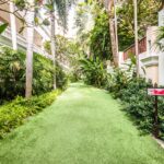 A serene pathway with lush green artificial grass flanked by various tropical plants. The area is lined with tall palm trees and shrubbery, leading towards a white building with railings. A "No Smoking" sign is visible on the right side of the pathway at the Grand Pacific Sovereign Resort & Spa.