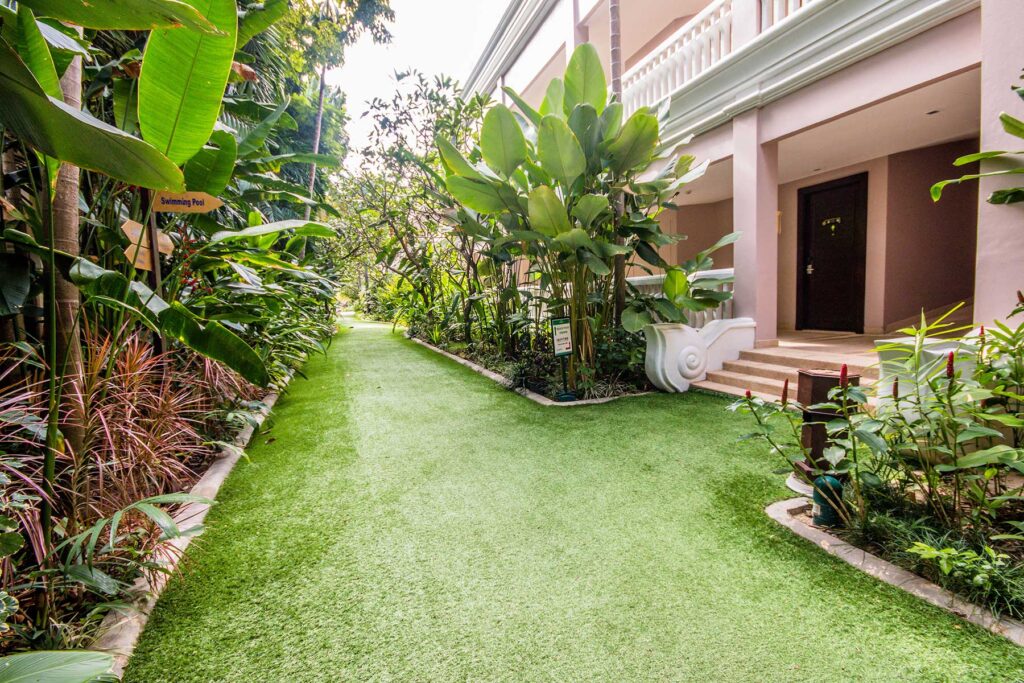 A lush, green pathway lined with tropical plants leads to the Grand Pacific Sovereign Resort & Spa entrance. The well-maintained artificial grass is bordered by a variety of leafy plants and shrubs, with a balcony featuring white railings and ornate stairs visible in the background – a prime off-market hotel investment opportunity.