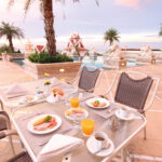 A luxurious outdoor dining area by the pool at the Grand Pacific Sovereign Resort & Spa, adorned with statues and palm trees. A table is set with plates of breakfast items, glasses of orange juice, and cups of coffee. The setting overlooks an ocean view with a clear sky during sunrise or sunset.