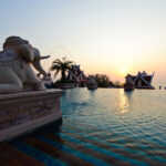 A serene outdoor pool at the Grand Pacific Sovereign Resort & Spa boasts water features shaped like elephants. The horizon displays a beautiful sunset, casting a golden glow across the water. Ornate buildings with pointed roofs are visible in the background, framed by palm trees—an exclusive off-market hotel investment opportunity.