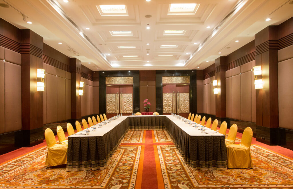 A spacious, well-lit conference room at the Grand Pacific Sovereign Resort & Spa features an elegant U-shaped table setup. The black and gold tablecloth is surrounded by yellow-gold chairs. With warm lighting, paneled walls, intricate decor, and a patterned carpet, it's the perfect off-market hotel investment opportunity.