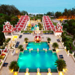The Grand Pacific Sovereign Resort & Spa is a luxurious seaside retreat, featuring red-roofed buildings encircling a large, illuminated central pool area. With multiple smaller pools and decorative landscaping by the ocean, this investment opportunity is nestled next to a serene forest.