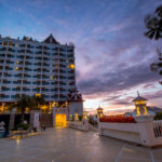 A tall hotel building with lit windows stands against a dramatic evening sky with hues of blue, purple, and pink. In the foreground, the spacious terrace of the Grand Pacific Sovereign Resort & Spa features outdoor seating and lantern-style lights. The scene exudes a serene and luxurious atmosphere.